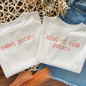 May include: Two light gray t-shirts with red text that reads "sweet potato" and "kind of rude potato". The shirts are folded and placed on a woven basket with decorative elements and blue jeans.