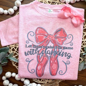 May include: Pink t-shirt with a gray and pink bow and ballet shoes. The shirt has the text "Let them praise His name with dancing" and "Psalm 149:3".