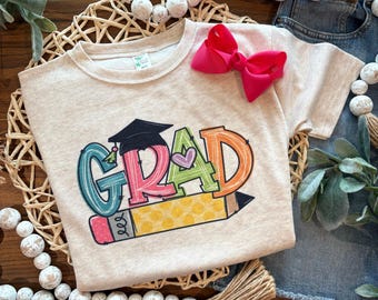May include: A light beige t-shirt with a colorful "GRAD" graphic, a graduation cap, and a pencil. The graphic includes teal, pink, orange, and green colors. A bright pink bow is attached to the shirt.
