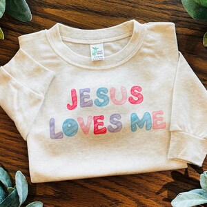 May include: A light gray long-sleeved shirt with the text "JESUS LOVES ME" in colorful, scribbled letters. The shirt is made of a soft material and is designed for children.