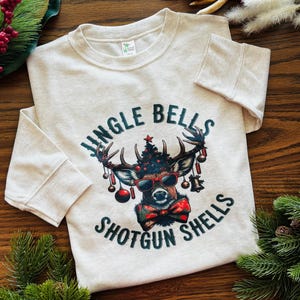 May include: A light beige sweatshirt with the text "Jingle Bells Shotgun Shells" in teal. The graphic features a deer wearing sunglasses, a bow tie, and a Christmas tree on its antlers. Red and green Christmas decorations are visible.