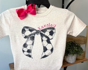 May include: A light gray t-shirt featuring a soccer ball bow design with the name "Kensley" in pink script. A bright pink bow is pinned to the shirt's neckline. The shirt is displayed on a black hanger.