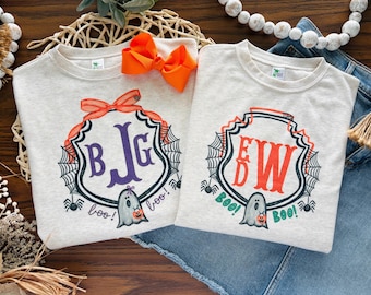 May include: Two light heather grey sweatshirts featuring a Halloween-themed graphic. Each shirt displays a black spiderweb crest with a ghost holding a pumpkin bucket, the text "boo!", and a personalised monogram in either purple or orange letters.