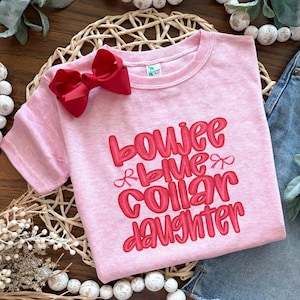May include: Pink t-shirt with the text "bowjee blue collar daughter" in red lettering. The shirt has a red bow on the front.