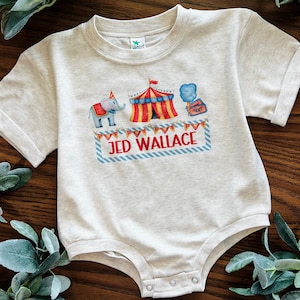 May include: A light gray baby bodysuit with rolled sleeves. The front features a circus-themed design with an elephant, a circus tent, and the name "JED WALLACE" in red letters. The design includes red, blue, and yellow colors.