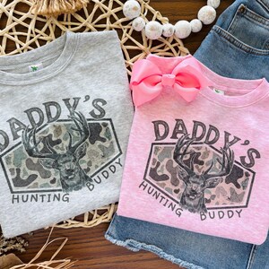 May include: A light gray sweatshirt and a light pink sweatshirt, each featuring a graphic print of a deer head inside a camouflage-patterned hexagon. The text "DADDY'S HUNTING BUDDY" is printed in dark letters around the central deer graphic.