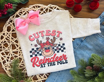 May include: A light beige sweatshirt with the text "Cutest Reindeer at the Pole" and a graphic of a reindeer with a red bow. A large pink bow is placed on the top left of the sweatshirt. The sweatshirt is placed on a woven mat with denim jeans.
