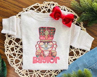 May include: A light gray long-sleeved sweatshirt featuring a colorful illustration of a nutcracker soldier with the name "Baylor" in red. A red bow is attached to the top of the sweatshirt. The sweatshirt is displayed on a woven placemat.