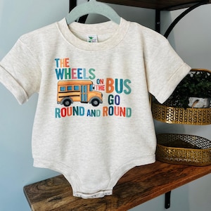 May include: A light beige baby onesie with rolled sleeves. The front features a colorful graphic of a yellow school bus and the text "THE WHEELS ON THE BUS GO ROUND AND ROUND" in various colors. The onesie is hanging on a light blue hanger.