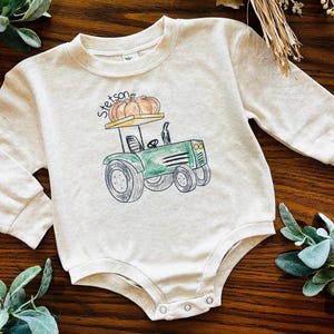 May include: A light beige baby bodysuit with long sleeves. The front features a green tractor graphic with a pumpkin on top and the word "Stetson" above it. The bodysuit has snap closures at the bottom.