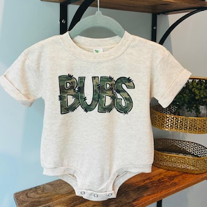 May include: A light beige baby onesie with short sleeves and the word "BUBS" in a camouflage print across the chest. The onesie has three snap closures at the bottom and is hanging on a gray hanger.