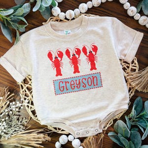 May include: A light beige baby onesie with three red lobster graphics. The name "Greyson" is printed in blue with a red dotted border. The onesie has short sleeves and a snap closure.