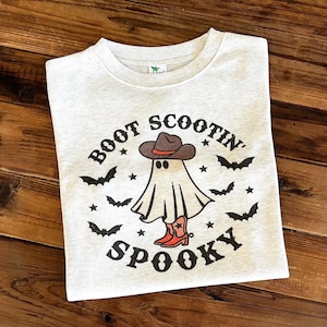 May include: A light gray t-shirt with a Halloween-themed graphic. The design features a ghost wearing a cowboy hat and boots, surrounded by bats and stars. The text reads "Boot Scootin' Spooky".