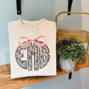 May include: A beige sweatshirt with a pink bow and a camouflage monogram design. The monogram features the letters "EMD" in a circular pattern. The sweatshirt is folded on a wooden shelf.