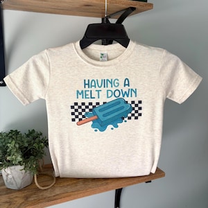 May include: A light beige t-shirt with the phrase "HAVING A MELT DOWN" in blue text. The shirt features a blue popsicle graphic with a melting effect and a black and white checkered pattern.