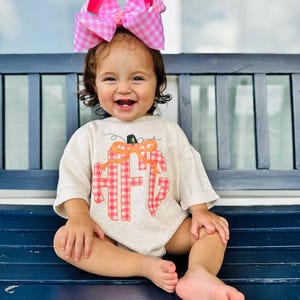 May include: A smiling toddler wearing a beige onesie with a pumpkin and monogram design. The onesie features a red and orange gingham pattern. The child is wearing a large pink and white gingham bow in their hair.