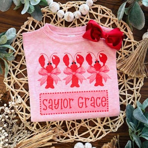 May include: Pink t-shirt featuring three red lobster illustrations with pink bows. The text "Saylor Grace" is printed in a pink rectangle. A red bow is placed at the top of the shirt.