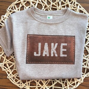 May include: A light gray t-shirt with the name "JAKE" printed on a brown leather-like patch. The shirt is made of a soft material and is displayed on a woven placemat. The shirt has short sleeves and a crew neck.