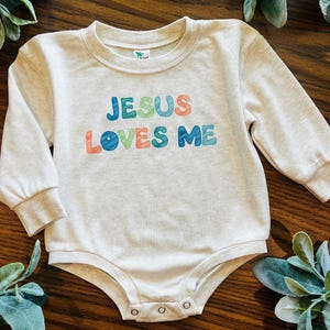 May include: A light gray baby bodysuit with long sleeves. The text "JESUS LOVES ME" is printed across the front in colorful letters. The bodysuit has a round neckline and snaps at the bottom.
