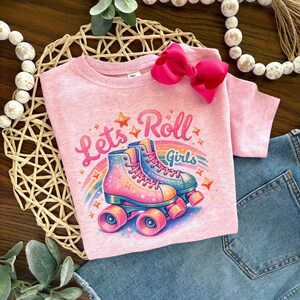 May include: Pink short-sleeved t-shirt with the text "Lets Roll Girls" and a graphic of colorful roller skates. A large pink bow is attached to the shirt. The shirt is paired with blue denim.