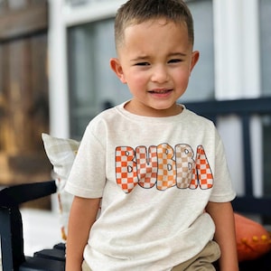 May include: A light beige t-shirt with the word "BUBBA" in a checkered orange and brown design. The t-shirt is short-sleeved and worn by a young child. The child is smiling and wearing khaki shorts.