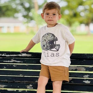 May include: A young child wearing a light beige t-shirt with a football helmet graphic and the name "lias" printed below. The child is also wearing brown shorts. The background is a blurred outdoor scene.