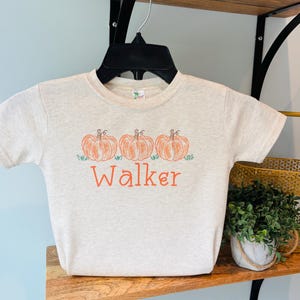 May include: A heathered beige short-sleeve t-shirt featuring a graphic of three orange pumpkins sitting on green grass. Below the pumpkins, the name Walker is printed in orange lettering. The shirt is displayed on a black plastic hanger.