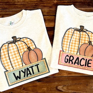 May include: Two off-white t-shirts featuring a graphic of two pumpkins, one with an orange gingham pattern and one solid orange. Below the pumpkins, a rectangular label displays the names WYATT on a blue-green background and GRACIE on a pink background.