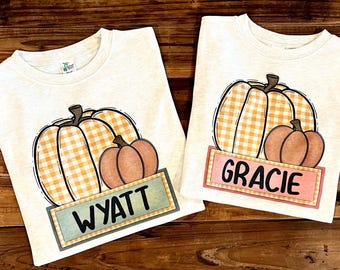 Puede incluir: Dos camisetas de color blanco roto con un gráfico de dos calabazas, una con estampado de cuadros naranja y otra naranja sólido. Debajo de las calabazas, una etiqueta rectangular muestra los nombres WYATT sobre fondo verde azulado y GRACIE sobre fondo rosa.