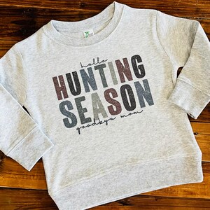 May include: A light gray sweatshirt with the words "hello HUNTING SEASON goodbye mom" printed in various colors. The sweatshirt has long sleeves and a crew neck, suitable for children.