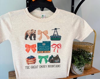 May include: A light beige t-shirt featuring colorful illustrations of a bear, bows, a cable car, pancakes, and a cabin. The text "THE GREAT SMOKY MOUNTAINS" is printed at the bottom. An orange bow is attached to the hanger.