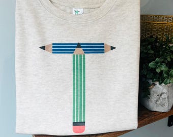 May include: A folded, light beige t-shirt featuring a graphic of a letter "T" made from pencils. The top pencil is blue with black tips, and the vertical pencil is green with a pink eraser.