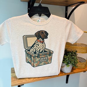 May include: A light gray t-shirt featuring a graphic of a dog inside a camouflage cooler with the text "USA". The dog has brown and white spots and is wearing an orange collar.