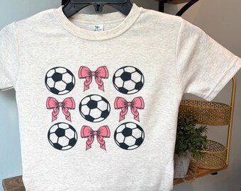 May include: A light gray t-shirt featuring a pattern of soccer balls and pink bows. The soccer balls are black and white, and the bows are a soft pink. The shirt is displayed on a black hanger.