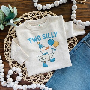 May include: A light beige long-sleeved shirt with the words "TWO SILLY" in blue. The shirt features a cartoon duck wearing a party hat, bow tie, and boots, holding blue and yellow balloons. The shirt is likely for a toddler's second birthday.