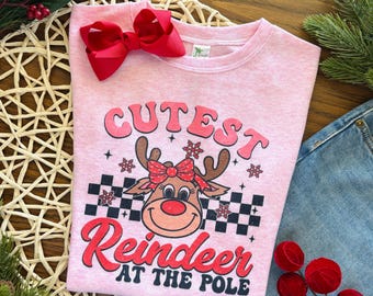 May include: A light pink t-shirt with the text "Cutest Reindeer at the Pole" and a cartoon reindeer graphic. A large red bow is pinned to the top of the shirt. The shirt is paired with denim shorts and Christmas decorations.