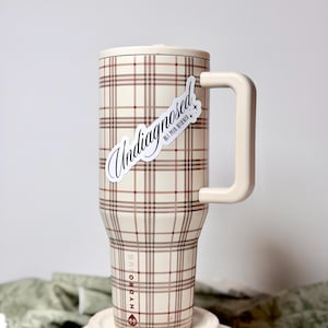 Può includere: Un Hydro Jug color crema con un motivo a quadri nei toni del marrone e del rosso. Ha un manico e un coperchio abbinati. Un adesivo con la scritta "Undiagnosed" è sul lato. Il logo Hydro Jug è visibile.
