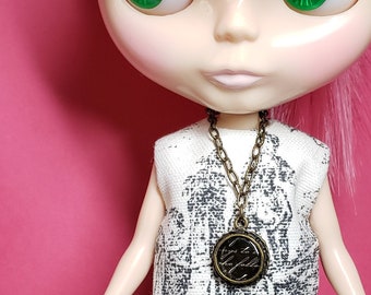 May include: A doll with pink hair and green eyes wearing a white and black patterned dress with a bronze chain necklace and a round pendant with the text "I try to be the fallen" on it.