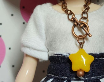 May include: A copper-toned chain necklace with a toggle clasp and a yellow star pendant. The star pendant has a small copper-colored bead at the bottom. The doll is wearing a white top and a denim skirt.