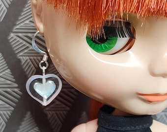 May include: A doll with red hair and green eyes wearing a silver heart-shaped earring with a clear iridescent center.