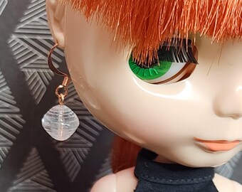 May include: A close-up of a doll wearing a clear, faceted crystal earring with a gold-colored hook. The doll has red hair, green eyes, and is wearing a black top. The earring is the focal point.