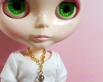 May include: A doll with green eyes and freckles wearing a white shirt and a gold chain necklace with a clear glass pendant.