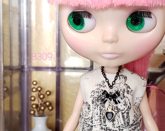 May include: A doll with pink hair and green eyes wearing a black and white patterned dress with a black necklace and a black bow charm with a black heart pendant. The doll is wearing a black necklace with a black bow charm and a black heart pendant.