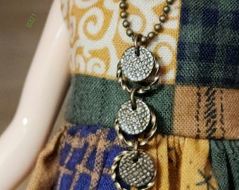 May include: A gold necklace with three round, textured, metal charms. The necklace is worn by a doll with a colorful patchwork dress.