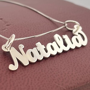 May include: A silver necklace featuring the name "Natalia" in a cursive script. The pendant hangs from a fine chain. The necklace is presented on a maroon surface.