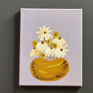 May include: A small canvas painting featuring a gold vase filled with white and yellow daisies. The background is a light purple color. The painting is a simple, yet elegant floral arrangement.