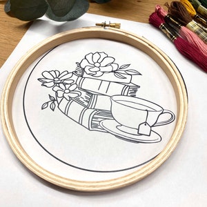 May include: A wooden embroidery hoop with a white fabric inside. The fabric has a black line drawing of a teacup, a stack of books, and flowers.