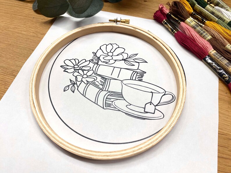 May include: A wooden embroidery hoop with a white fabric inside. The fabric has a black line drawing of a teacup, a stack of books, and flowers.