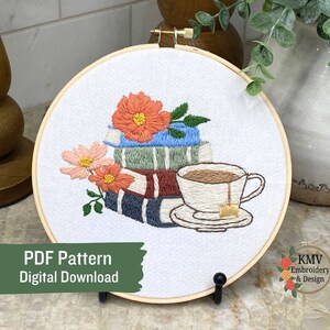 May include: Embroidered hoop art featuring a stack of books with flowers and a cup of tea. The design is in shades of blue, green, red, and brown. The text "PDF Pattern Digital Download" is at the bottom of the image.