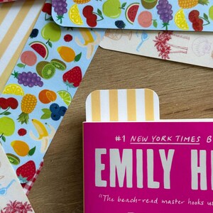 May include: A collection of colorful bookmarks with fruit and striped patterns. One bookmark is inserted into a pink book with the text "#1 NEW YORK TIMES B EMILY HI". Other bookmarks feature illustrations of fruits like strawberries, bananas, and grapes.